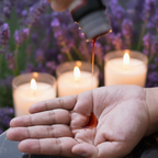 Hand pouring liquid from a bottle into another hand with candles and lavender in the background